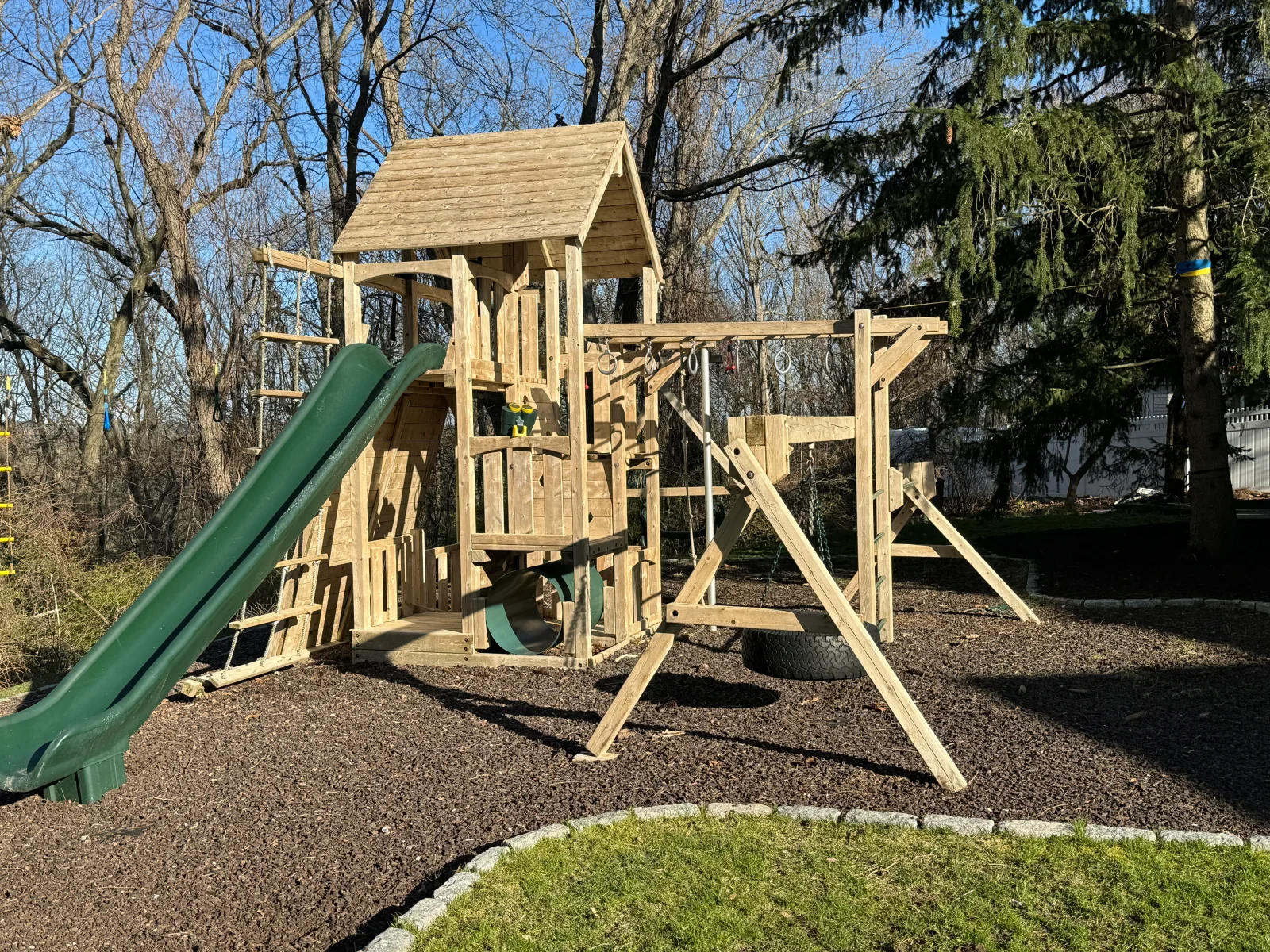 Wooden playset with slide, climbing features, and peaked roof structure on mulch ground